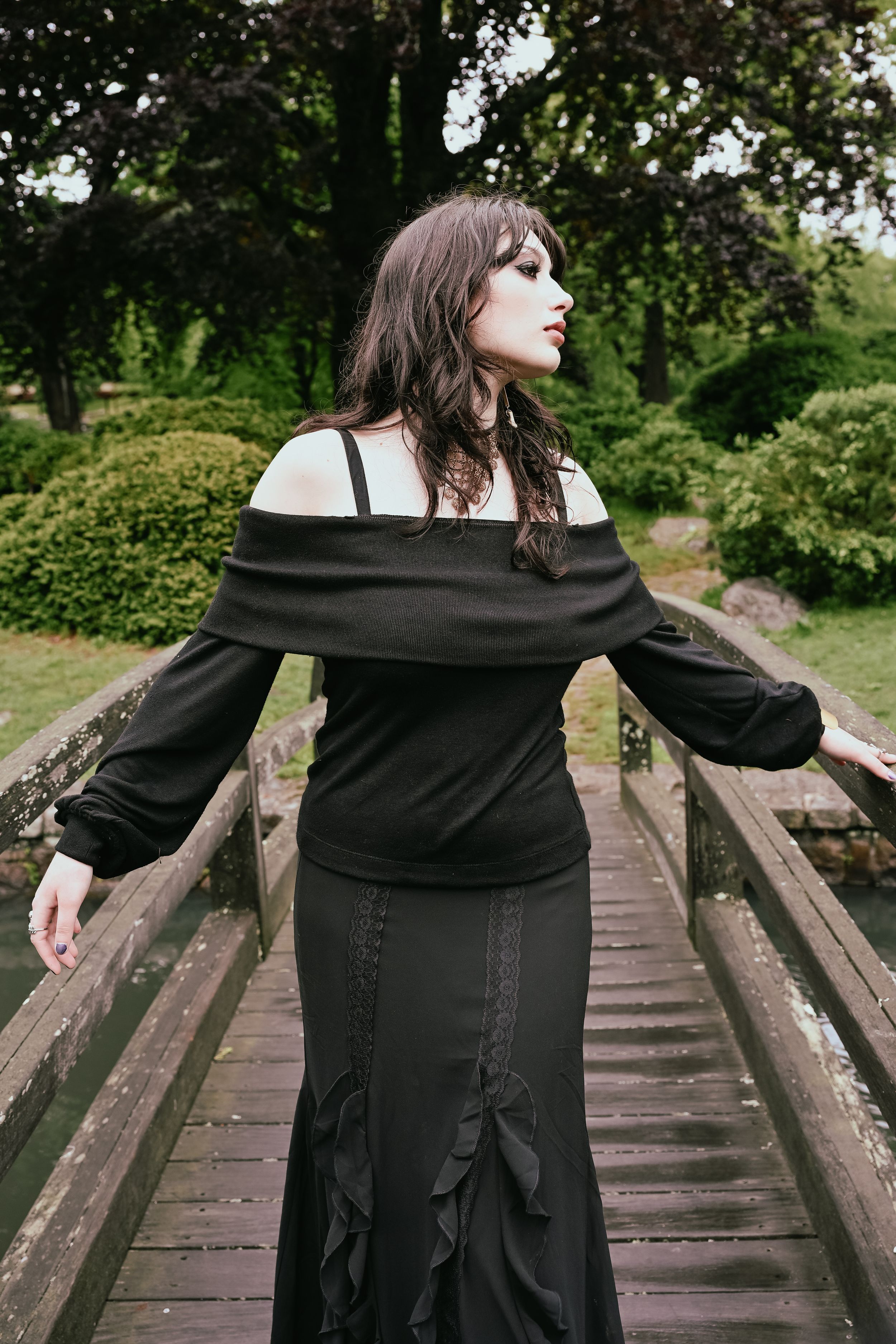 Gothic woman in black off-shoulder dress with arms outstretched on a wooden bridge in a lush garden.