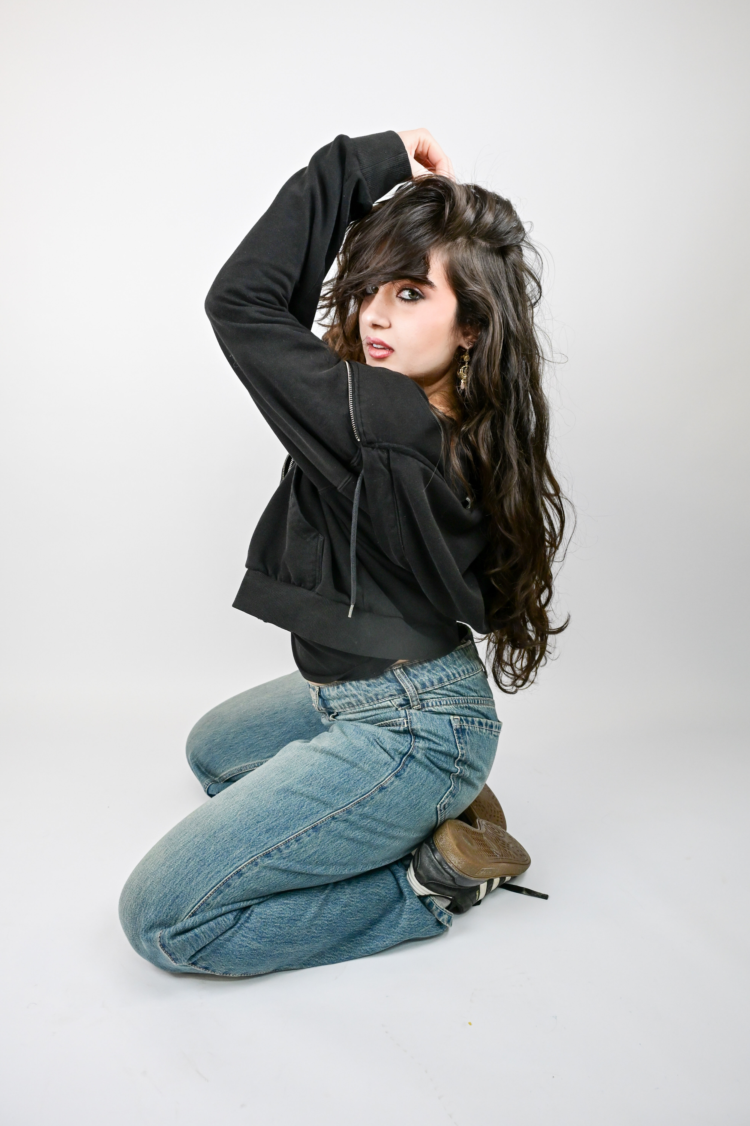 Brunette woman kneeling in jeans and black jacket with long wavy hair in a studio setting.