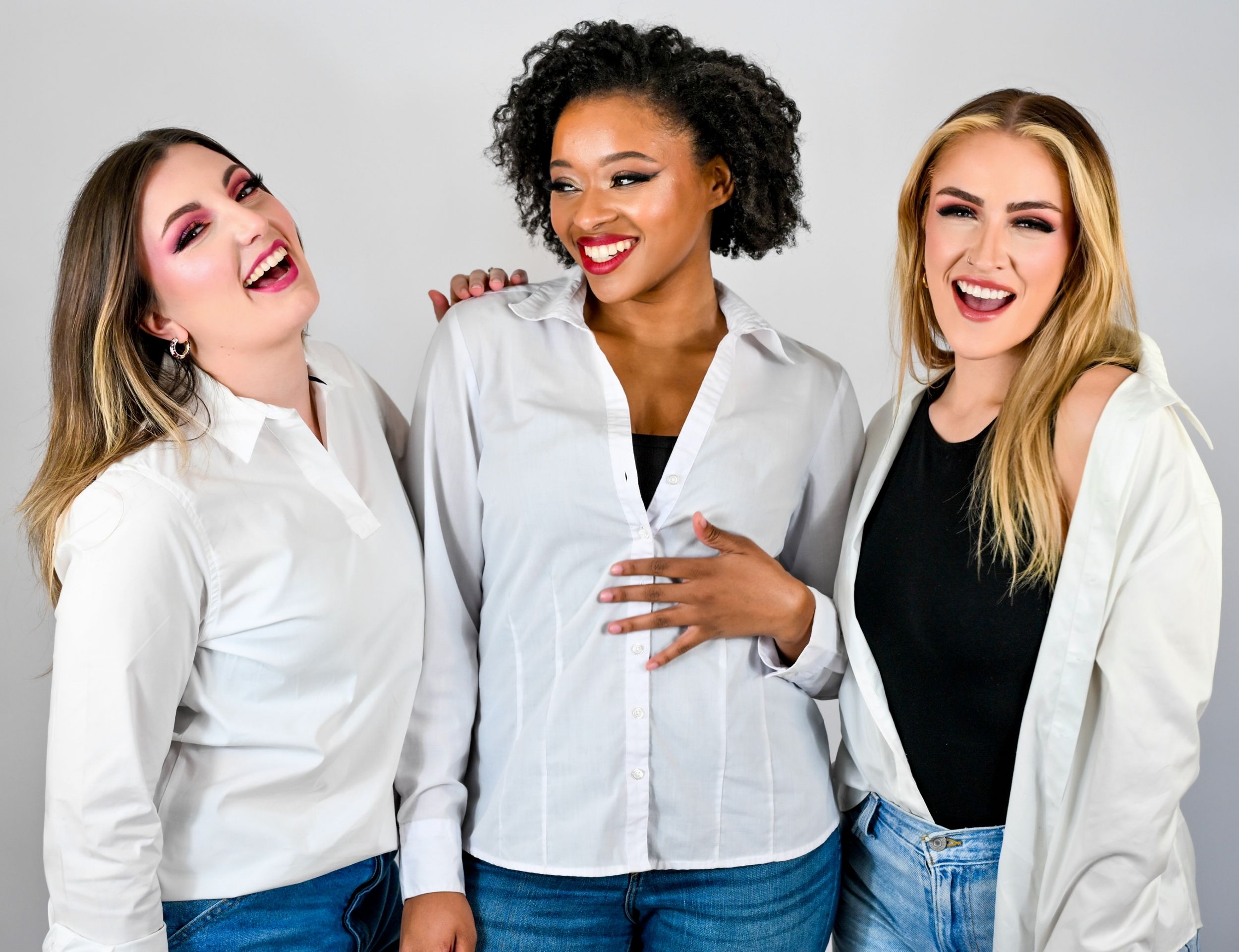 Three diverse women laughing and posing together in white shirts and jeans against a light background.
