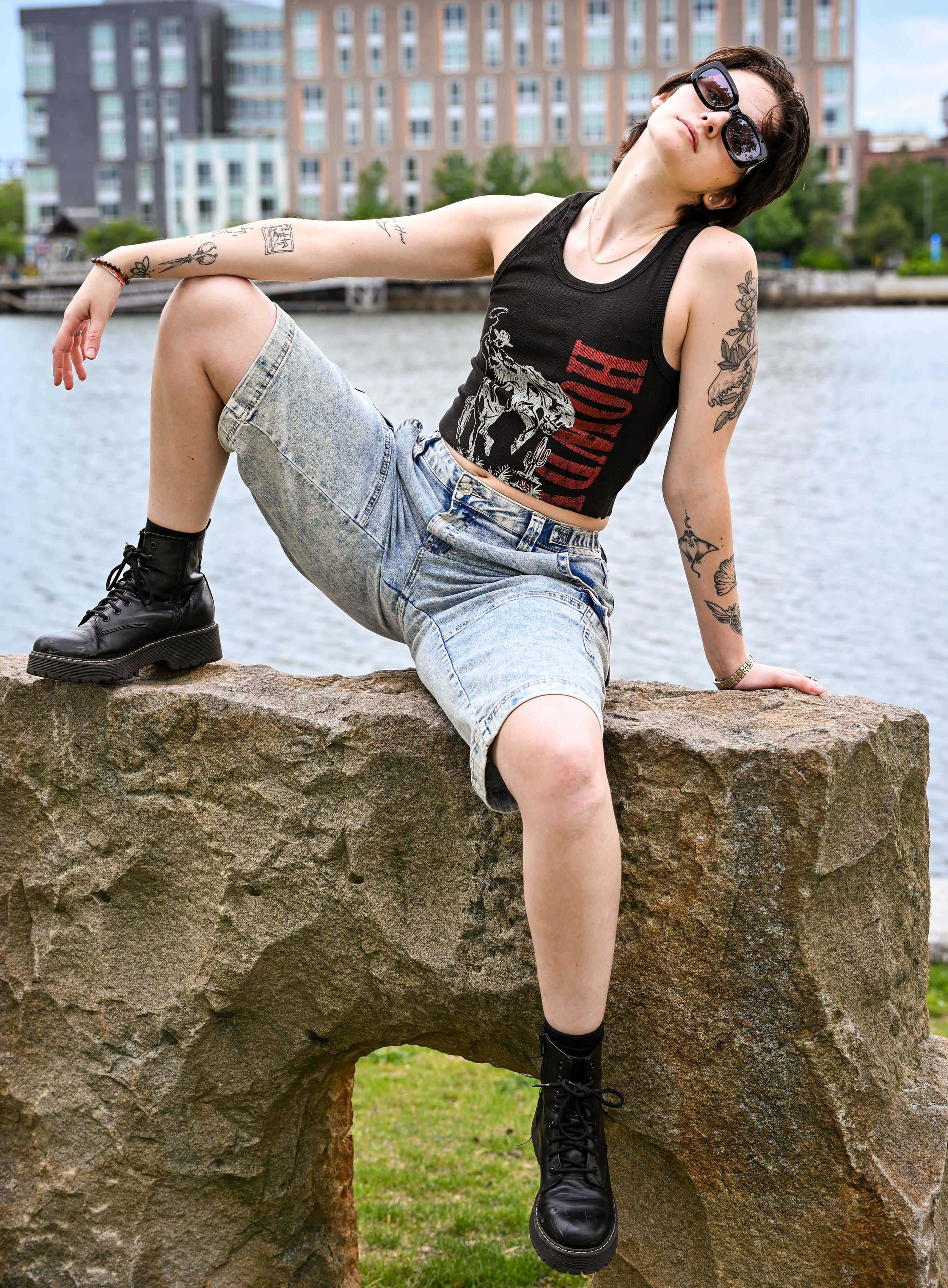 Tattooed woman in black crop top, denim skirt, and combat boots posing on rocks near waterfront.