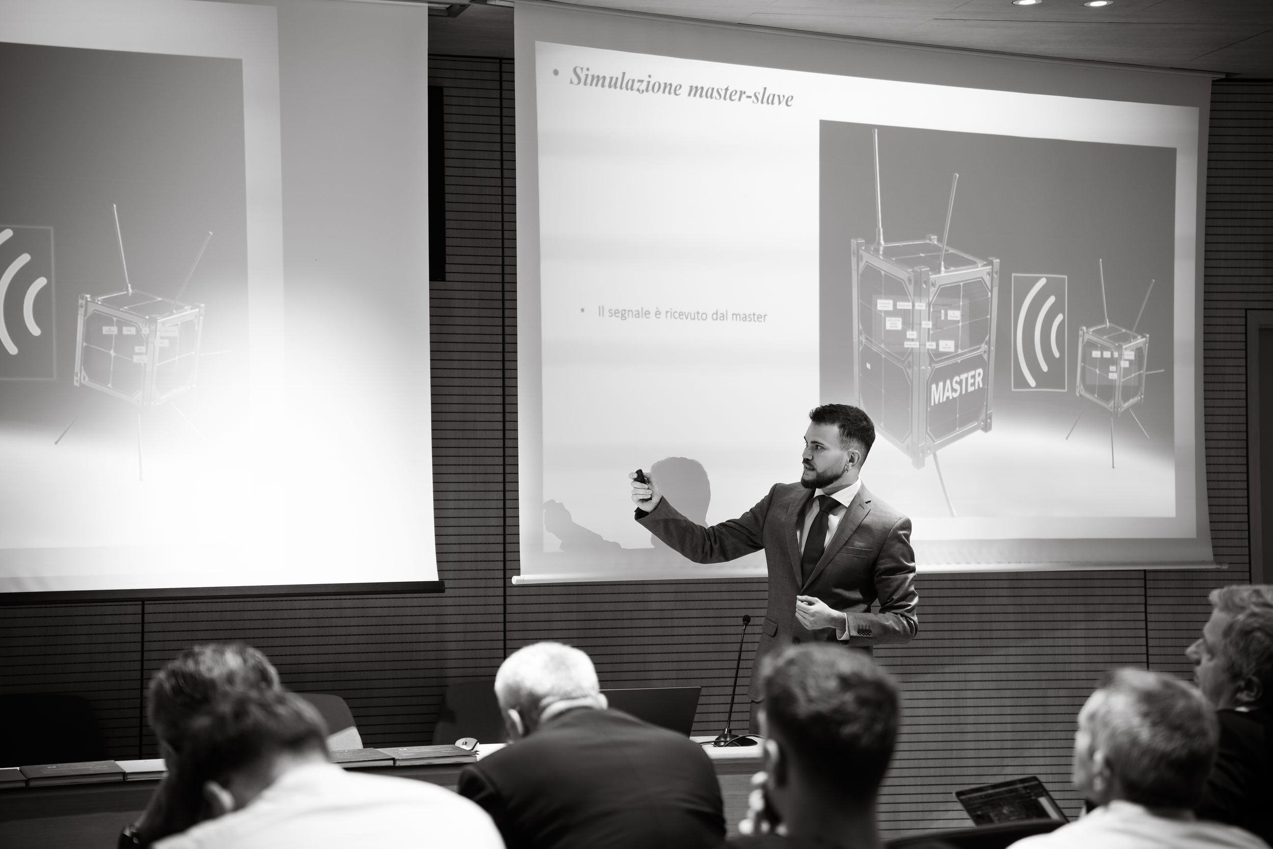 A man in a suit presents a slideshow to an audience in a darkened conference room, black and white photo.
