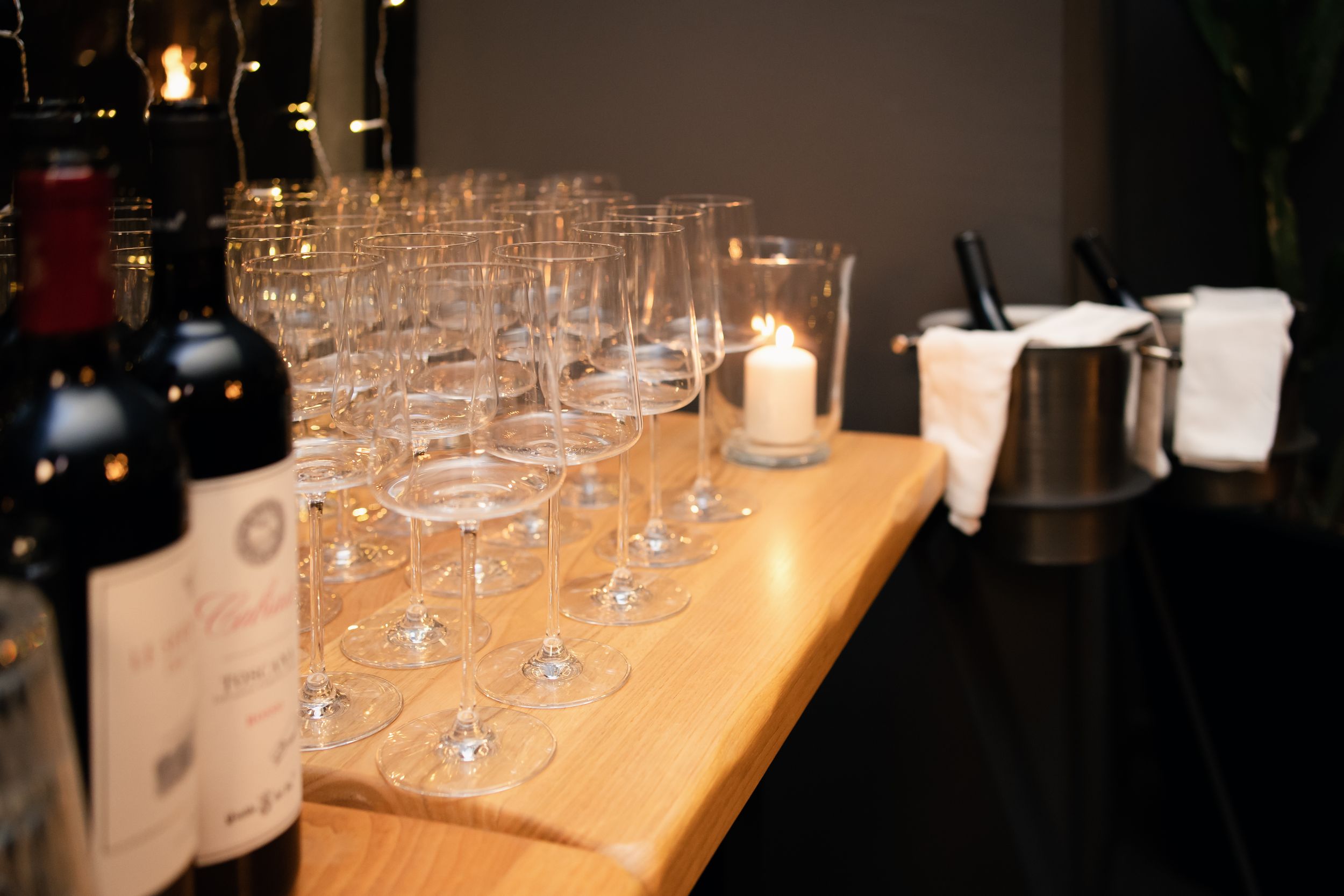 Elegant wine glasses lined up on a wooden bar counter with a lit candle and wine bottles at an upscale event.