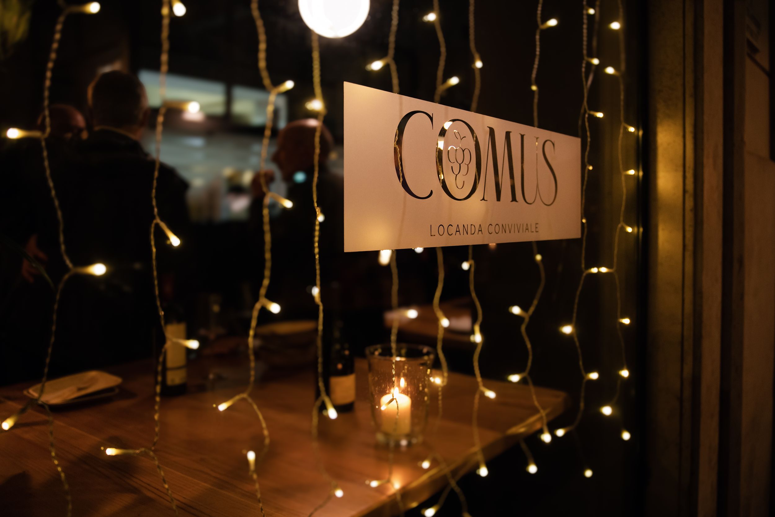 Comus Locanda Contadina restaurant sign glowing warmly amid fairy lights with candlelit wooden table in cozy dark interior.