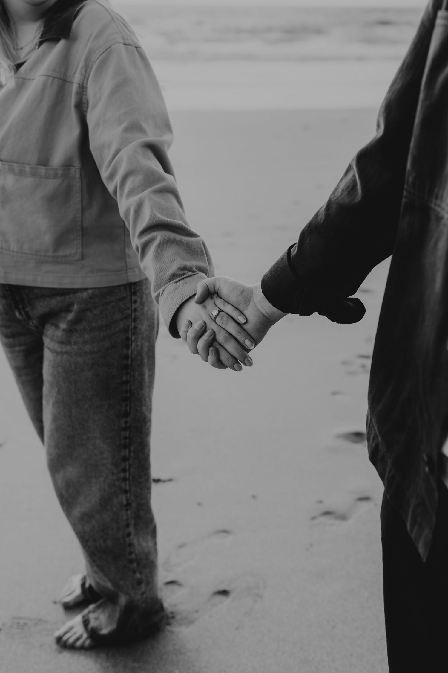 Black and white photo of a couple holding hands on a sandy beach, one person barefoot, wearing jeans and a light jacket.