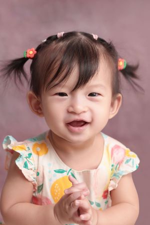 Smiling Asian toddler girl with pigtails and floral dress clapping hands against pink background.