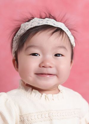 Smiling Asian baby girl wearing a white lace headband and cream knit sweater against a pink background.