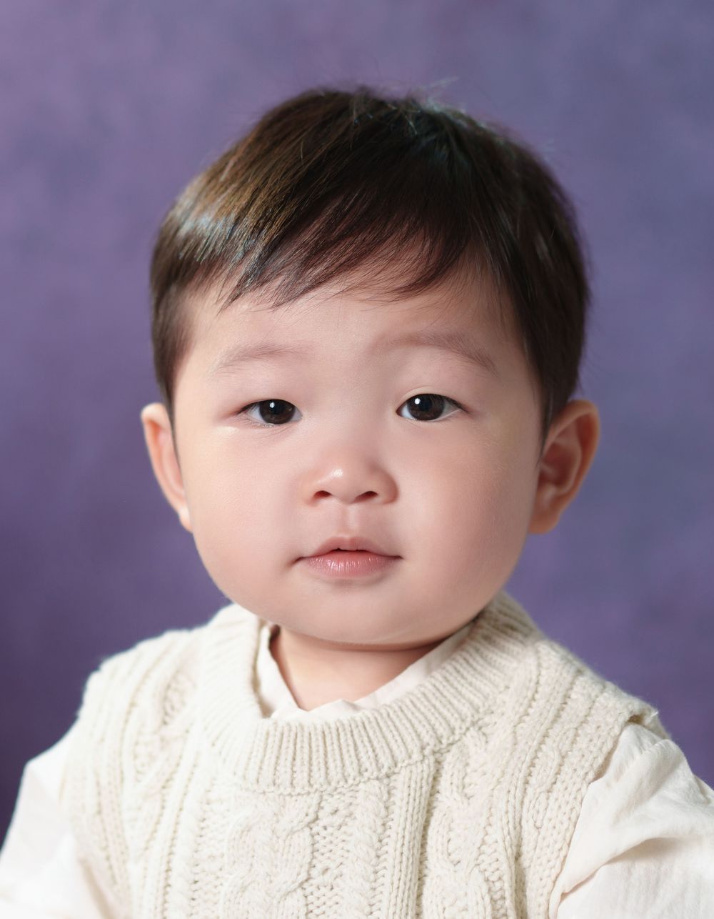 Adorable Asian toddler in a white cable-knit sweater poses for a portrait photo against a purple background.