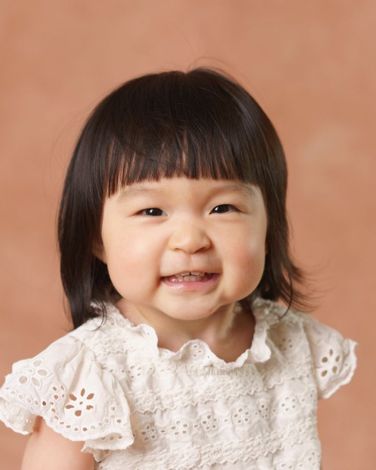 Smiling toddler girl with dark hair and bangs wearing a white eyelet lace dress against a warm beige background.