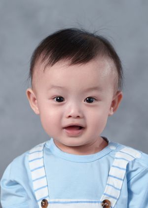 Smiling Asian baby boy in blue striped shirt with suspenders, portrait photo on gray background.