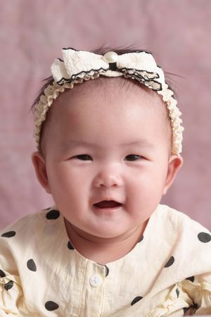 Smiling Asian baby girl wearing a cream polka dot outfit and decorative bow headband against a pink background.