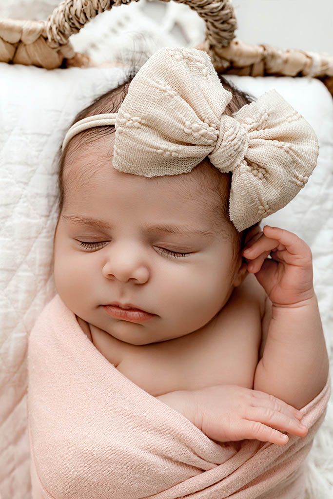 Sleeping newborn baby girl wrapped in pink swaddle with cream bow headband in wicker basket.