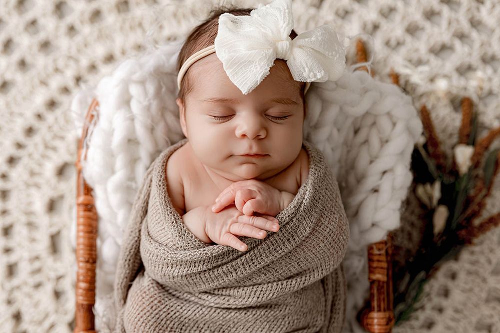 Sleeping newborn baby girl swaddled in taupe wrap with white bow headband in wicker basket.