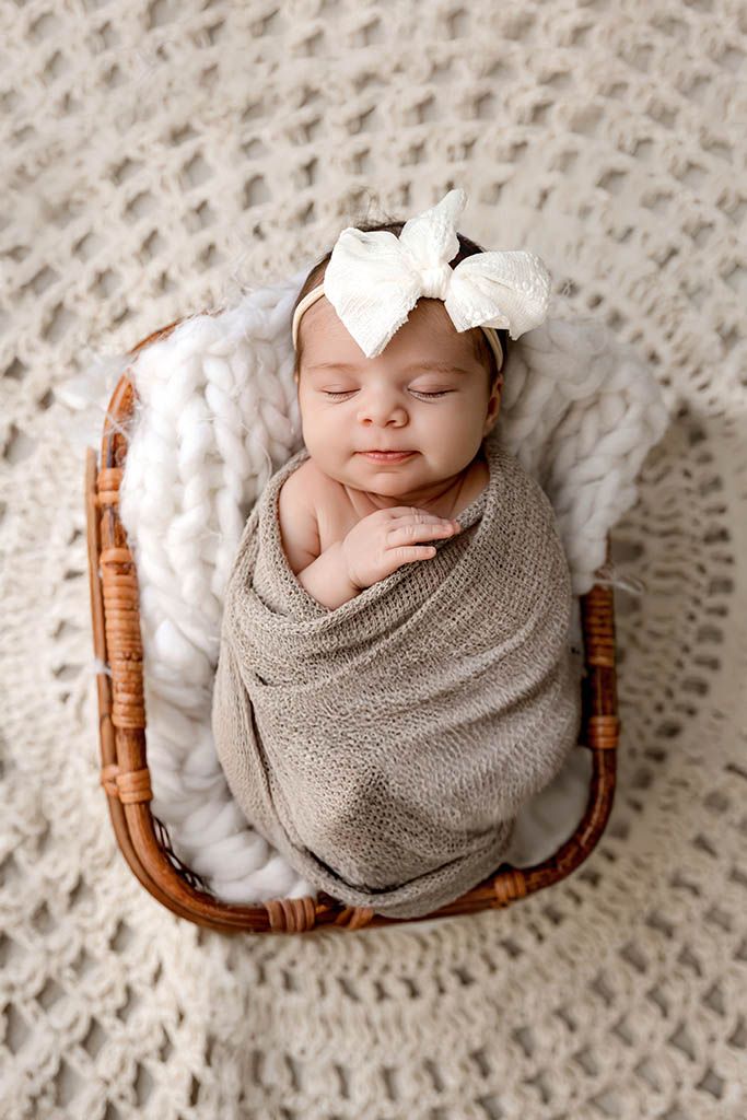 Smiling newborn baby girl swaddled in taupe wrap with white bow headband sleeping in wicker basket.