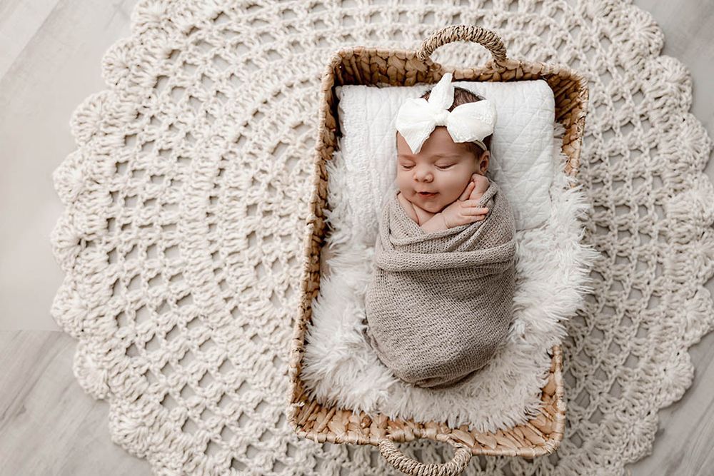 Newborn baby girl swaddled in cream wrap with white bow headband sleeping in wicker basket.