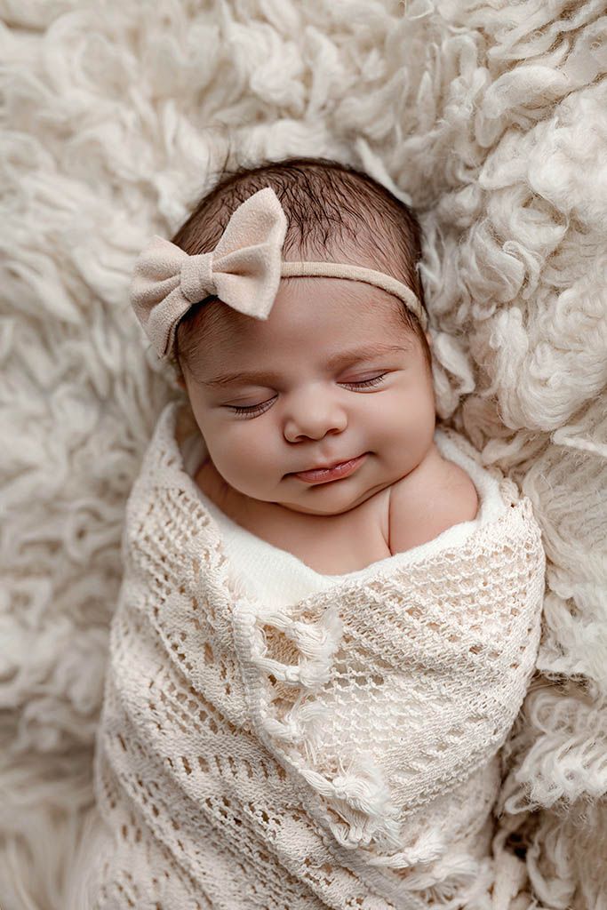 Peaceful newborn baby girl wrapped in ivory knit swaddle with beige bow headband sleeping on fluffy rug.