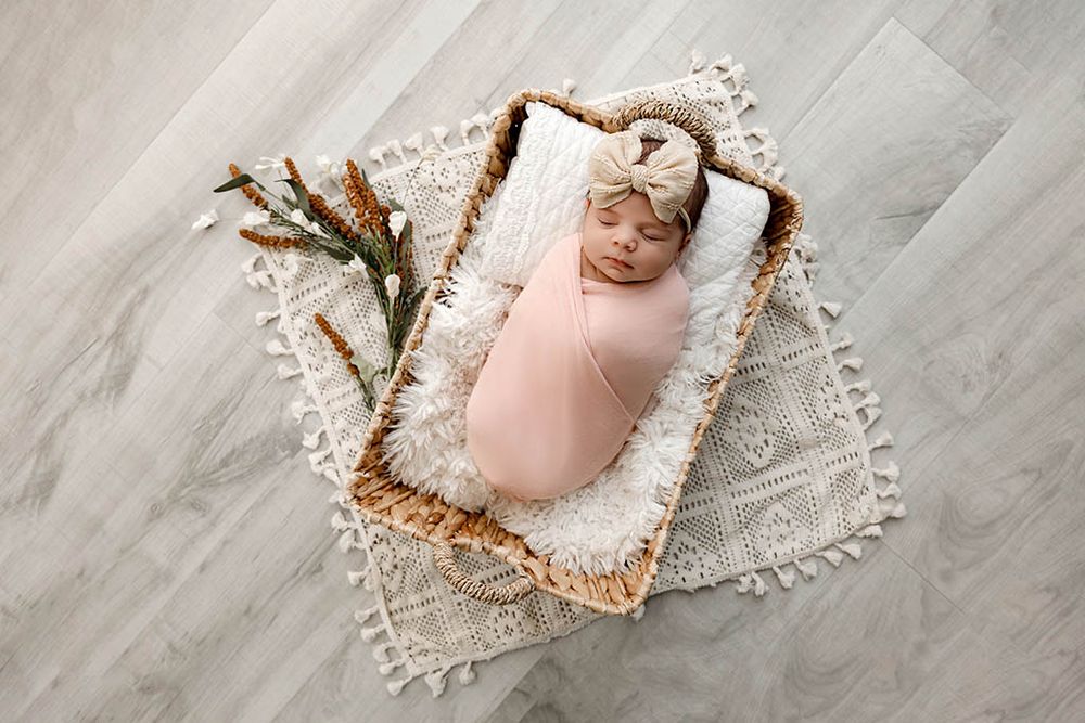 Winchester CA newborn photographer photo of baby girl in pink swaddle and bow headband sleeping peacefully in wicker basket with greenery.