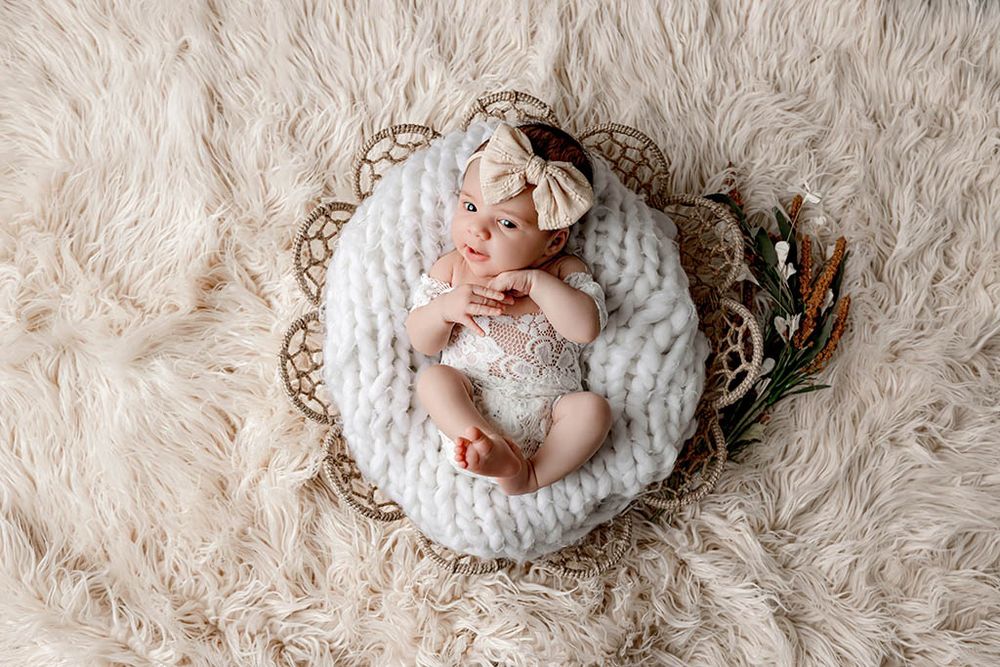 Newborn baby sleeping in a wicker basket with chunky knit blanket and bow headband on fluffy cream rug.