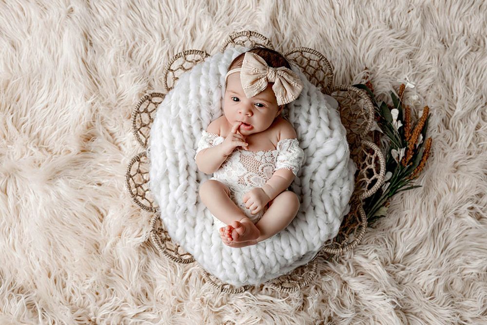 Newborn baby girl in white lace romper and bow headband sleeping in a wicker basket with chunky knit blanket.