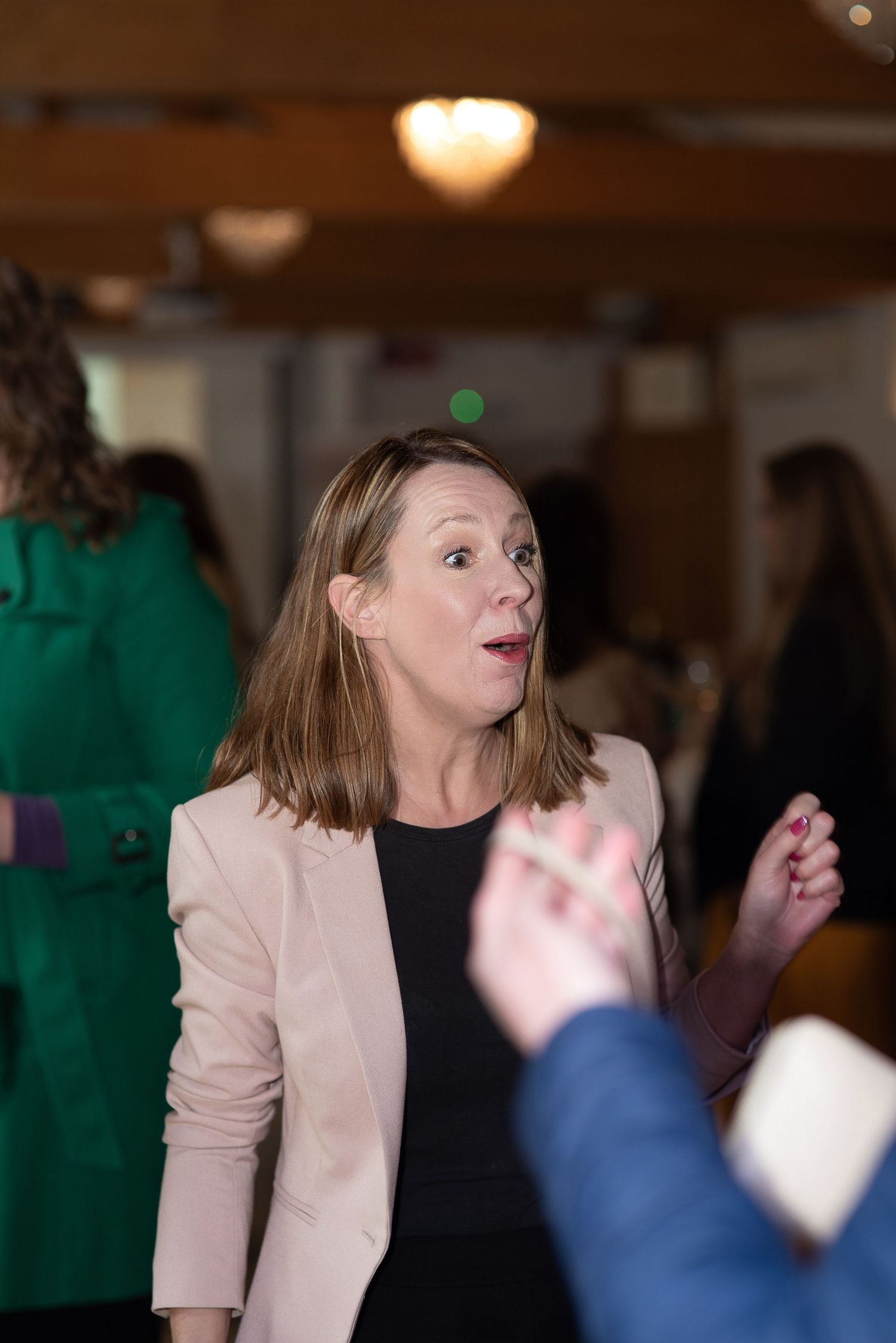 Woman in pink blazer and black top conversing at a networking event with blurred attendees in background.