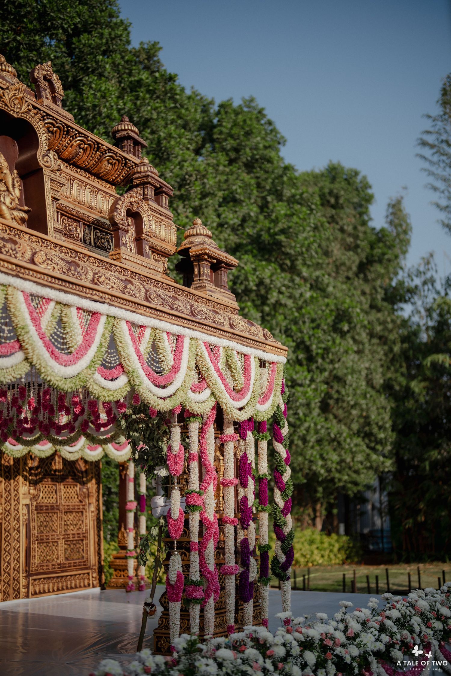 Ornate Indian wedding mandap adorned with vibrant floral garlands in pink, white, and green against lush trees.