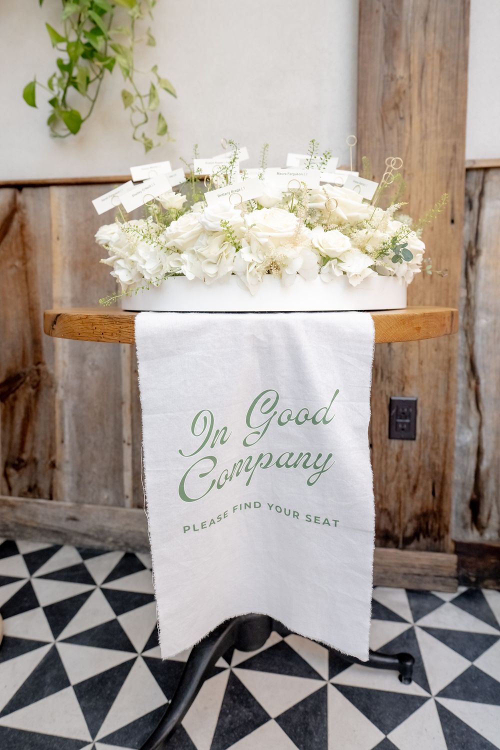 Elegant wedding seating chart display with white floral arrangement and 'In Good Company' banner on black and white tile floor.