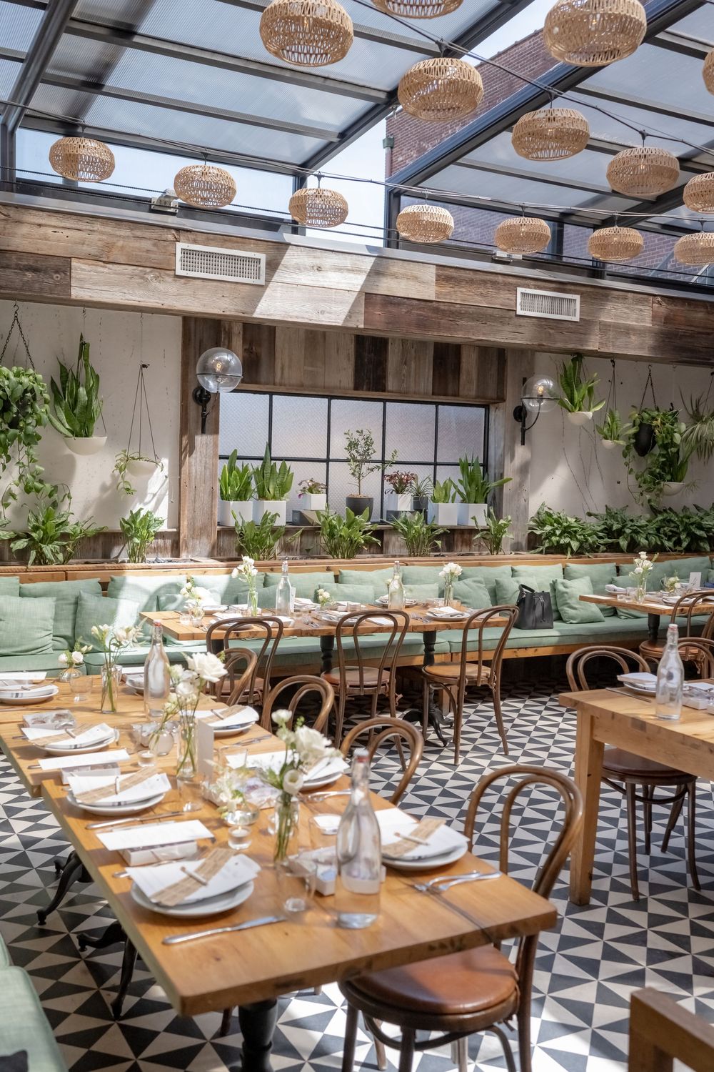 Bright restaurant interior with wooden tables, bentwood chairs, green banquettes, patterned tile floors, and hanging rattan lights.