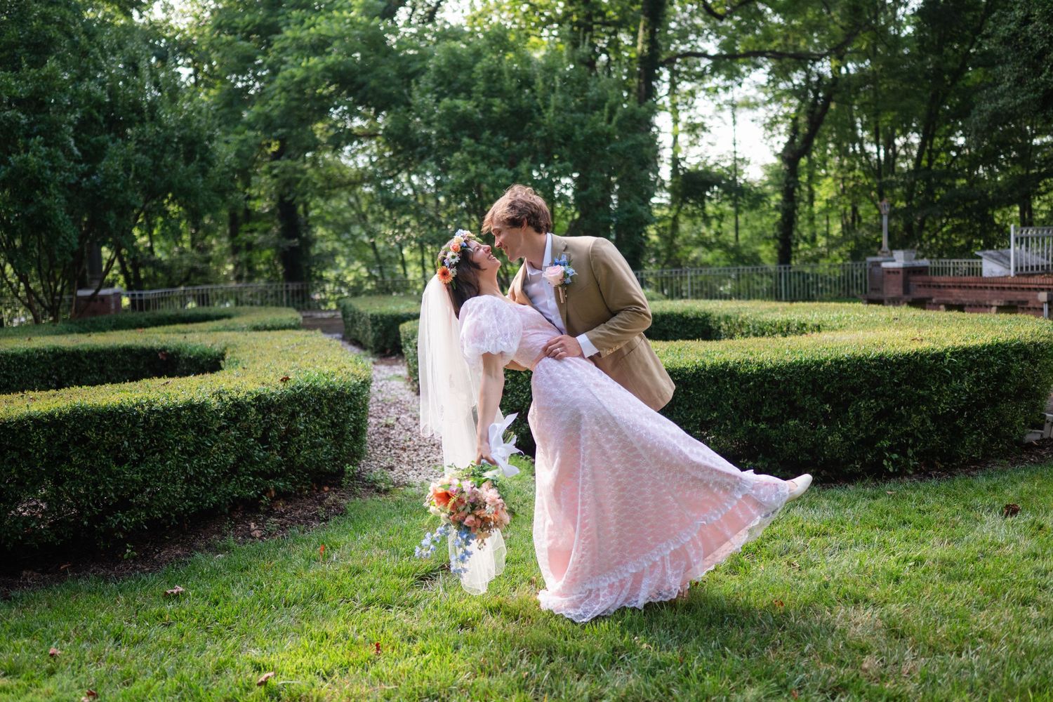 Groom dips bride in lush garden setting, bride holding colorful bouquet in elegant white wedding dress.