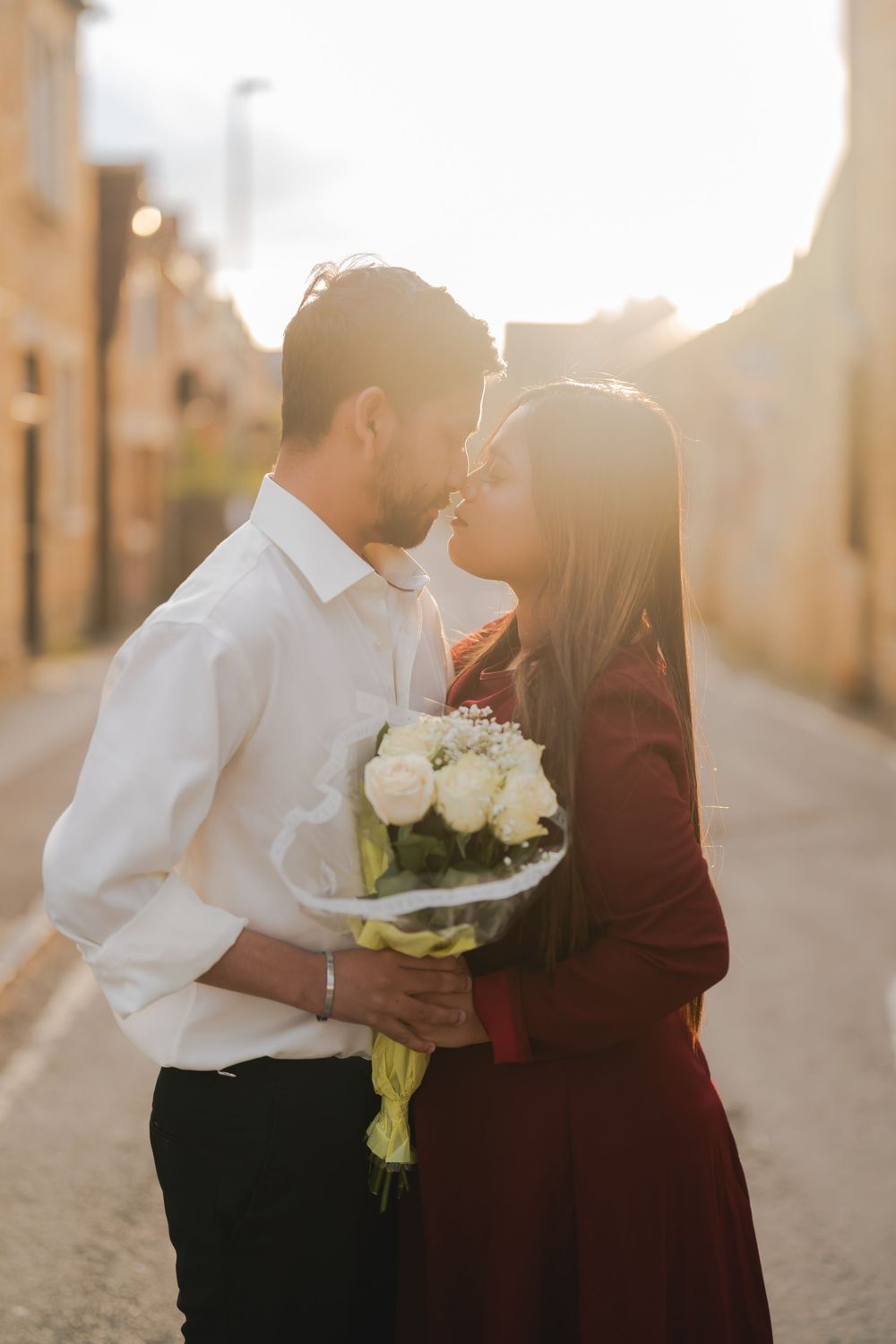 Romantic couple sharing a kiss on a sunlit street, man holding a bouquet of white roses.