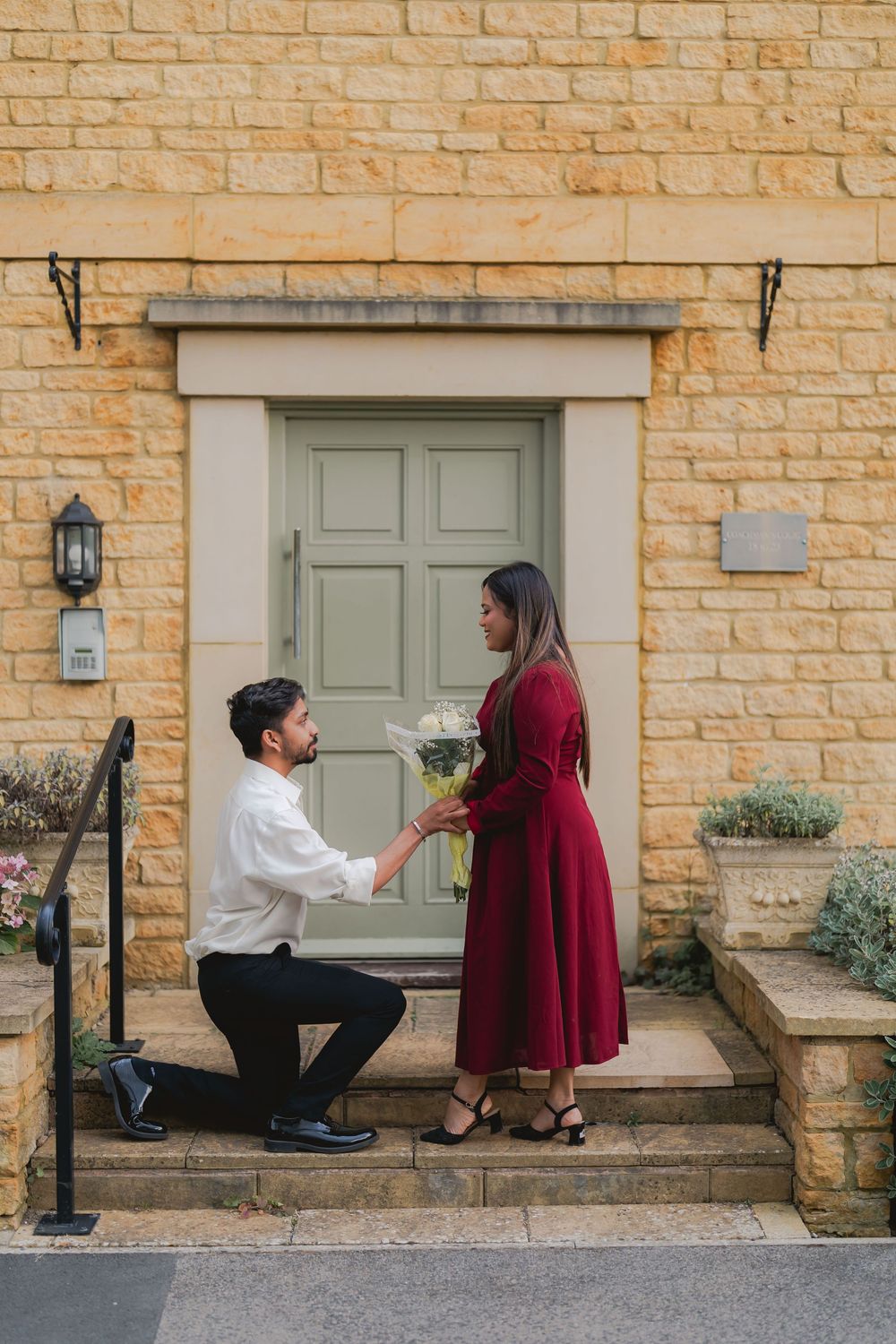 Man on one knee proposing to woman in red dress on steps of stone building with green door.