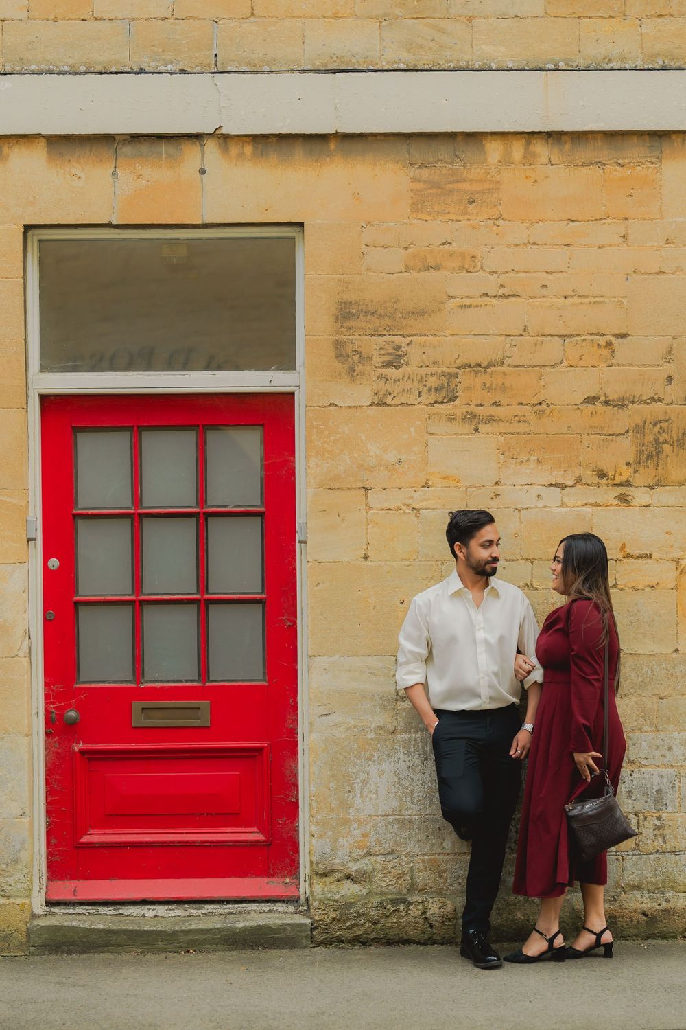 Couple standing by a vibrant red door against a golden stone wall, woman in burgundy dress, man in white shirt.