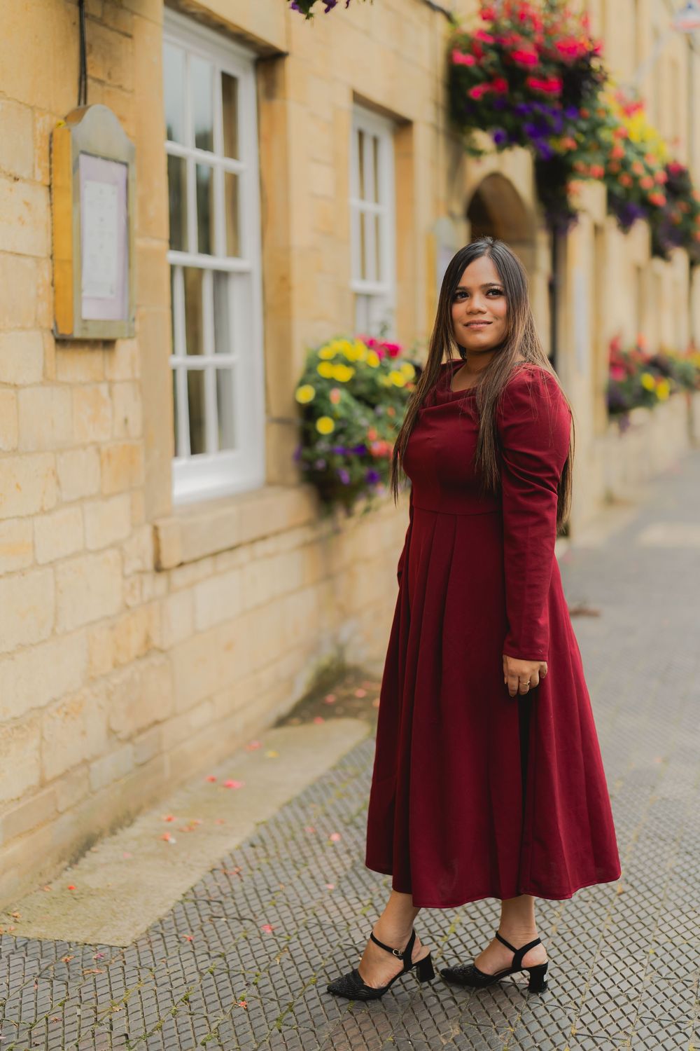 Woman in burgundy midi dress and black heels posing on cobblestone street beside stone building with flower displays.