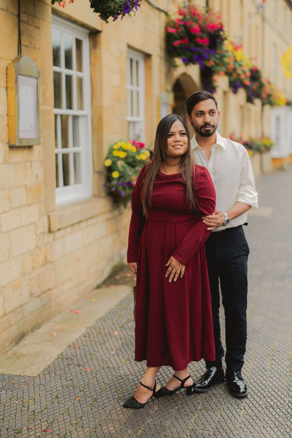 Couple posing in front of a stone building with colorful hanging flowers, woman in burgundy dress, man in white shirt.