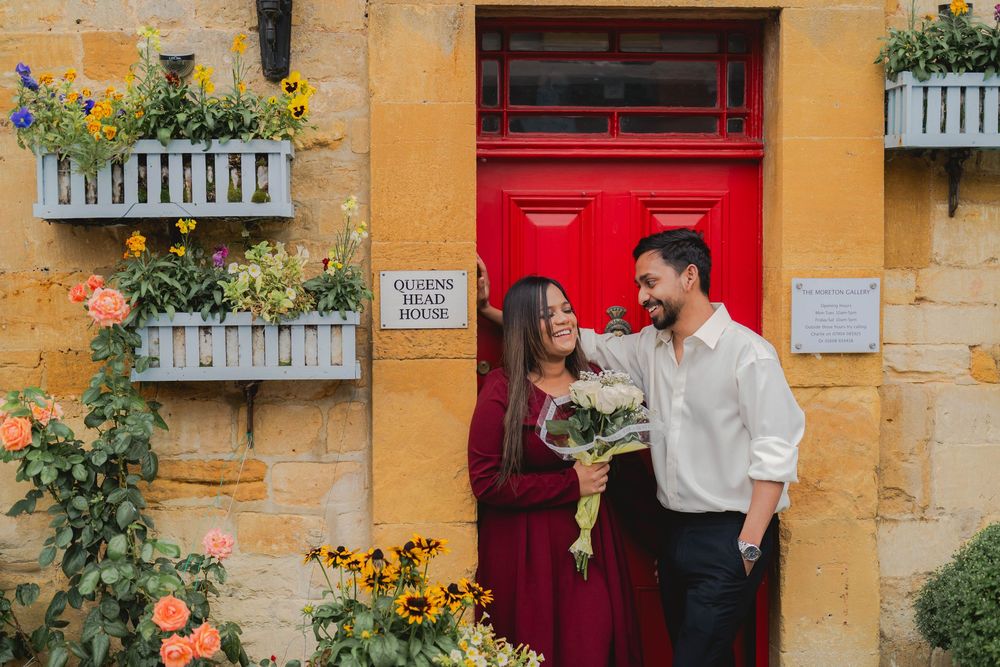 Couple smiling and holding flowers outside a charming stone building with a red door and colorful blooms.