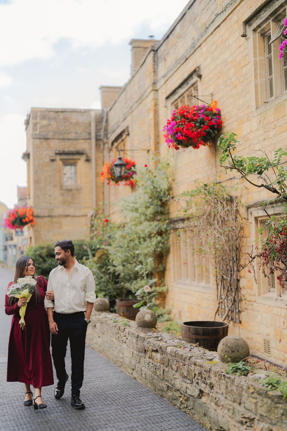 Couple strolling past a charming stone building adorned with vibrant hanging flower baskets in a quaint English town.