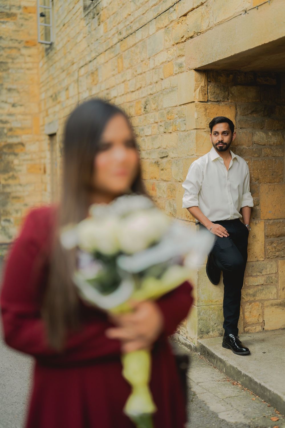 A man in a white shirt leans against a stone wall while a woman in red holds white roses in the foreground.