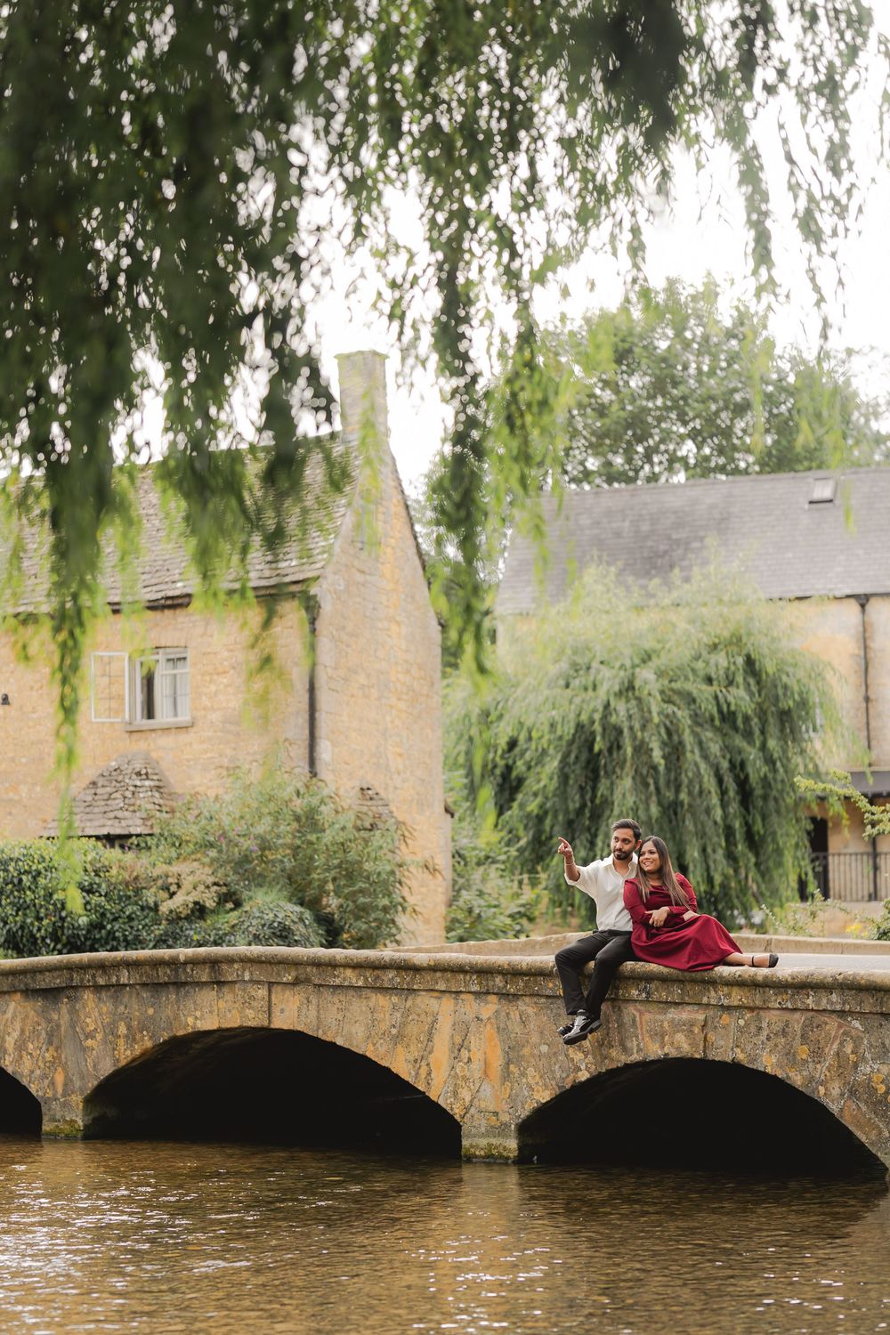 Couple sitting on a stone bridge over a river beside a charming Cotswolds village with lush greenery.