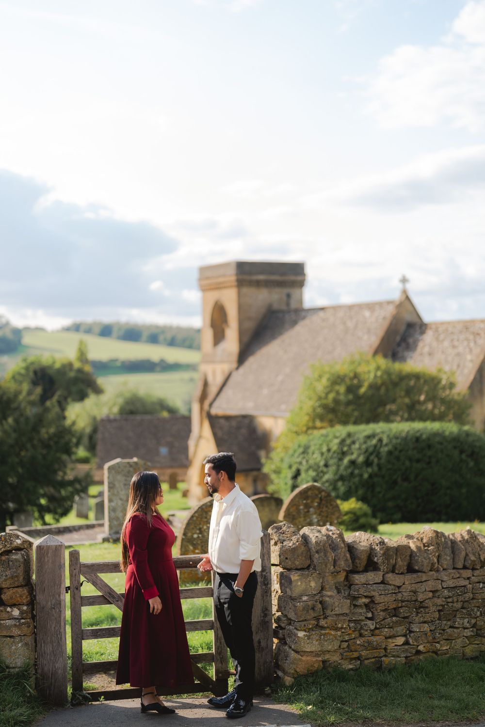 Couple in romantic attire standing by a stone gate with a historic English church and green hills behind them.