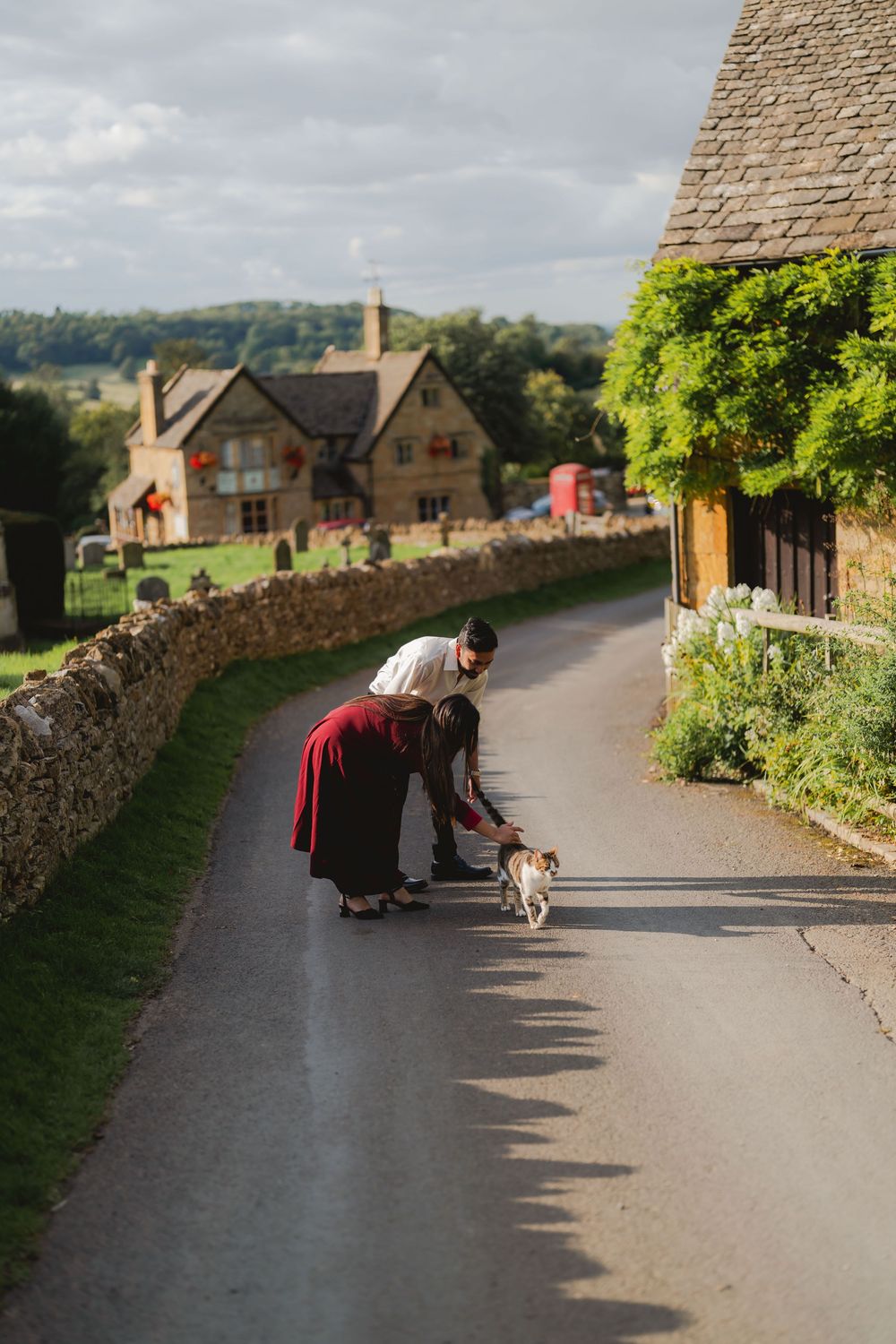Couple bending down to pet a small dog on a quiet countryside village road lined with stone walls and traditional cottages.