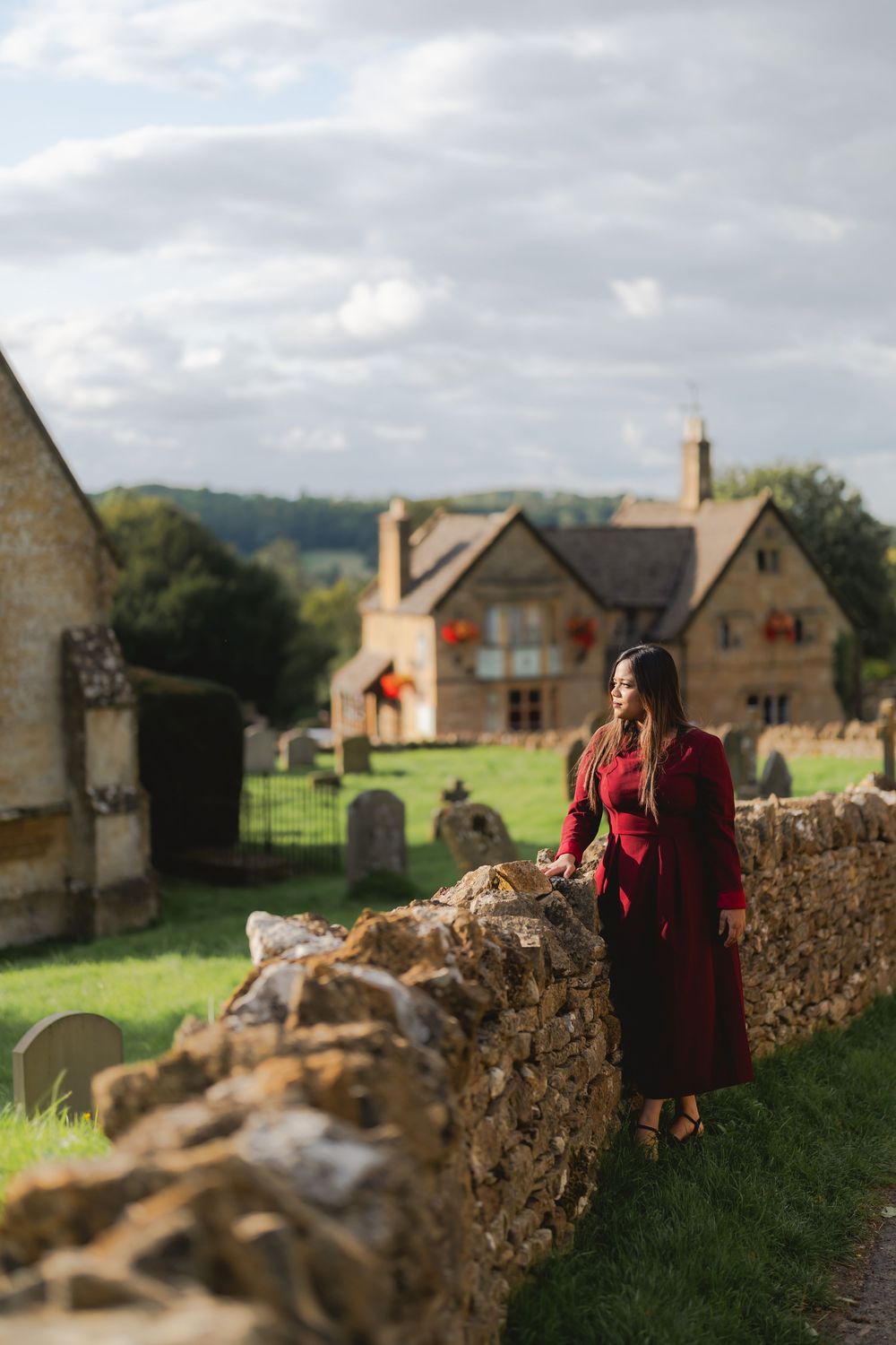 Woman in red coat stands by stone wall in English Cotswolds village with historic church and manor house backdrop.