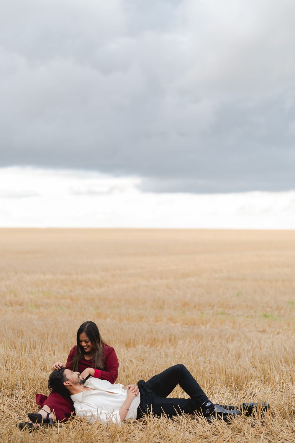 Couple relaxing in a golden wheat field under dramatic cloudy skies, woman sitting beside reclining man.
