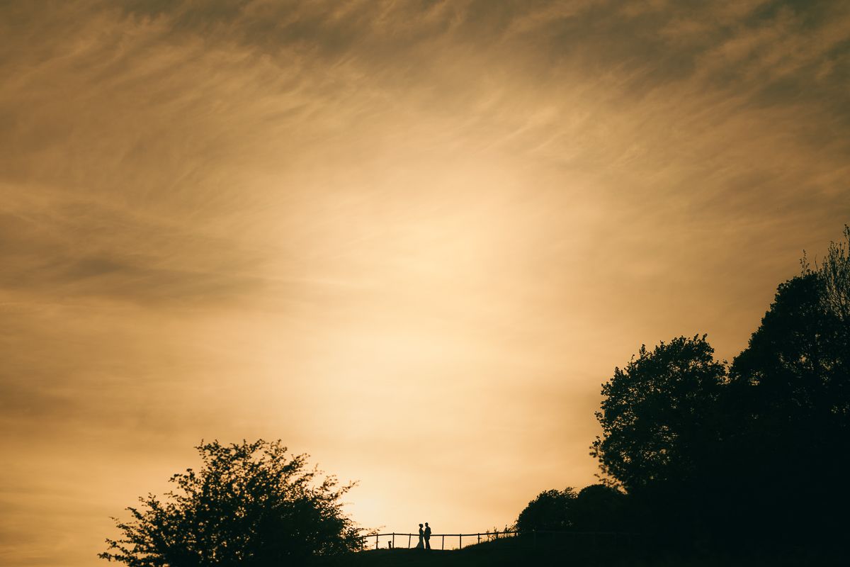 Silhouetted couple on a bridge at sunset with golden sky and trees in the background.