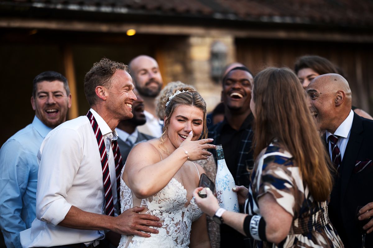 Joyful wedding couple laughing with guests outdoors, bride in lace gown and groom in burgundy tie.