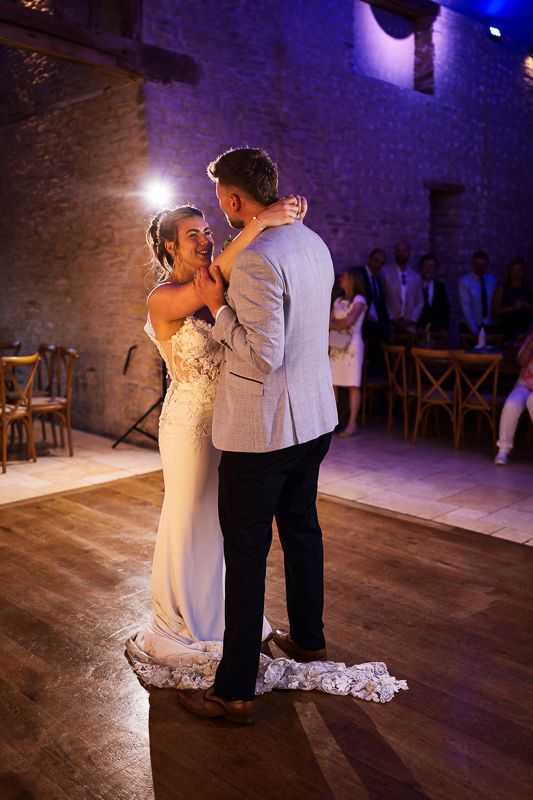 Bride and groom share their first dance in a rustic stone venue, bride in a fitted lace gown smiling joyfully.
