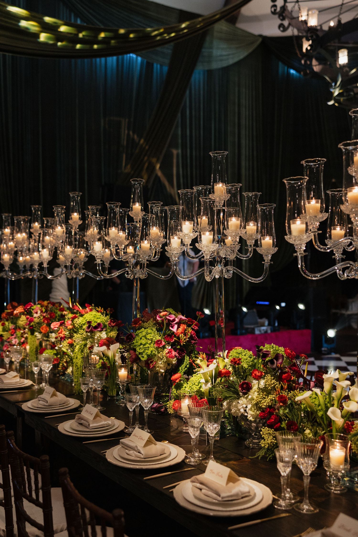 Elegant wedding reception table with crystal candelabras, white candles, red and green floral centerpieces, and formal place settings.