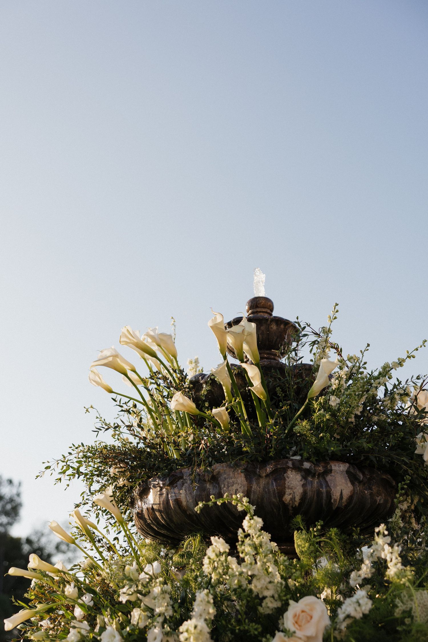 Stone fountain decorated with white calla lilies, roses, and lush greenery against a clear blue sky.