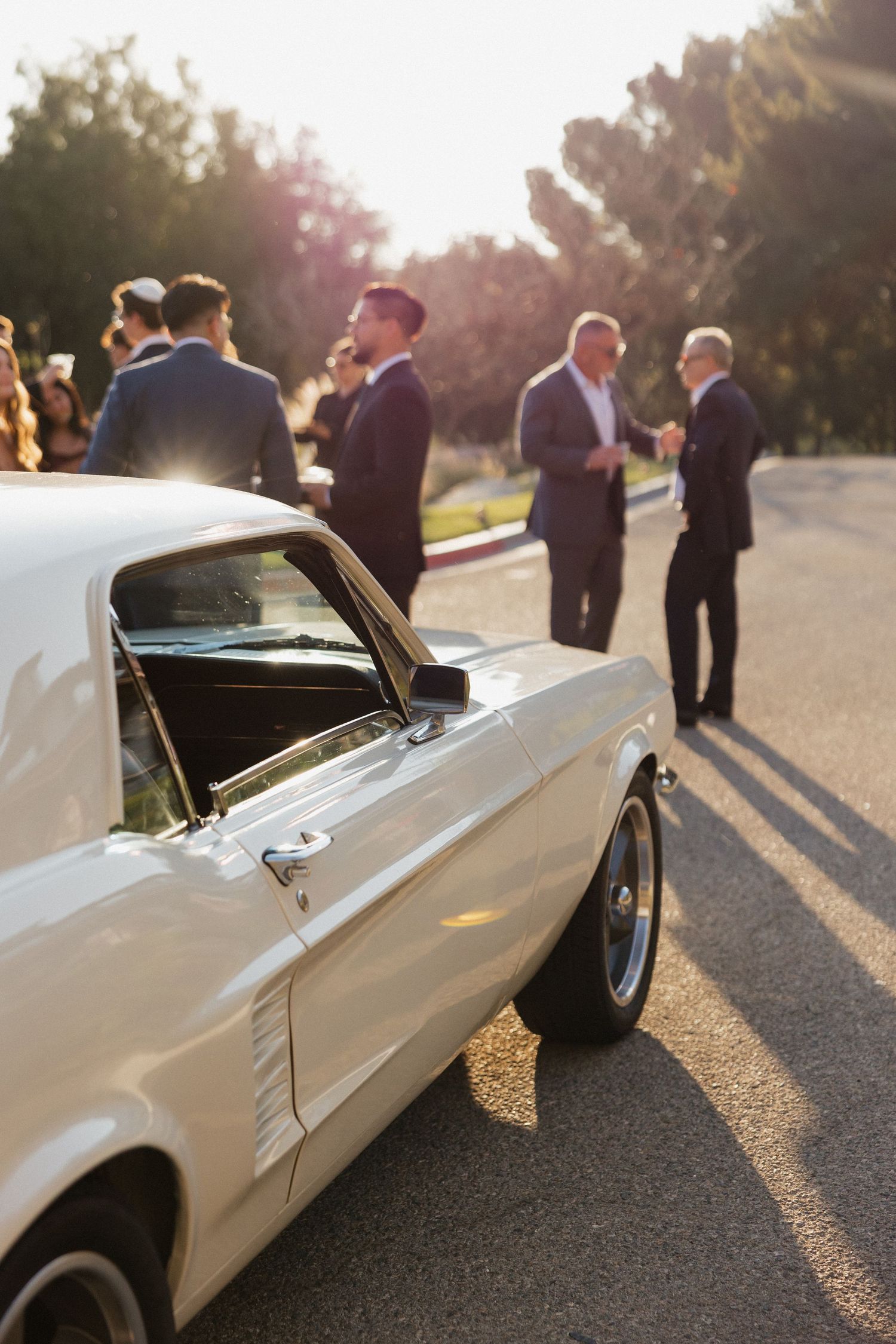 White vintage Ford Mustang parked outside sunny wedding venue with guests mingling in background.