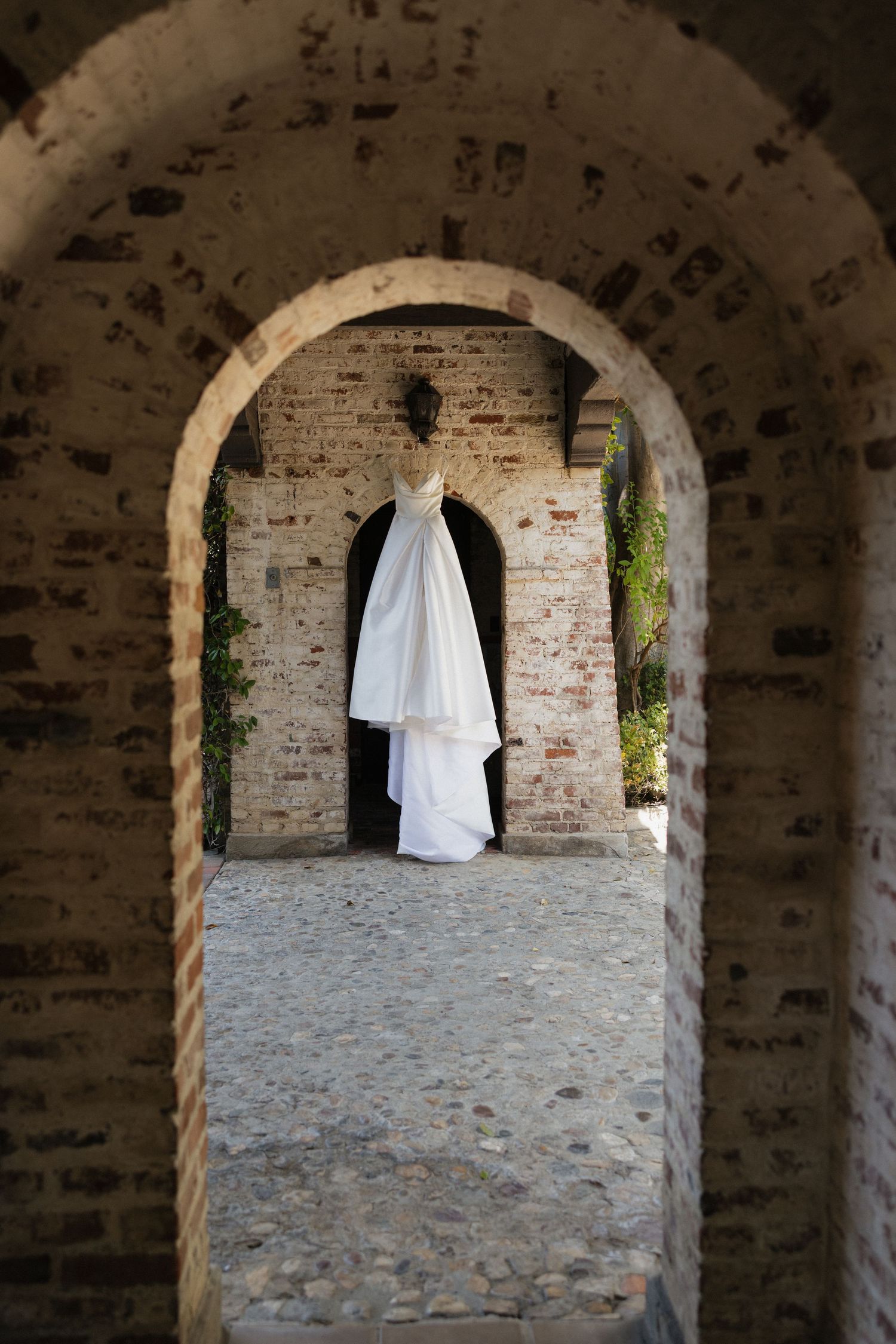 White wedding dress hanging in a rustic stone archway corridor with cobblestone floor.