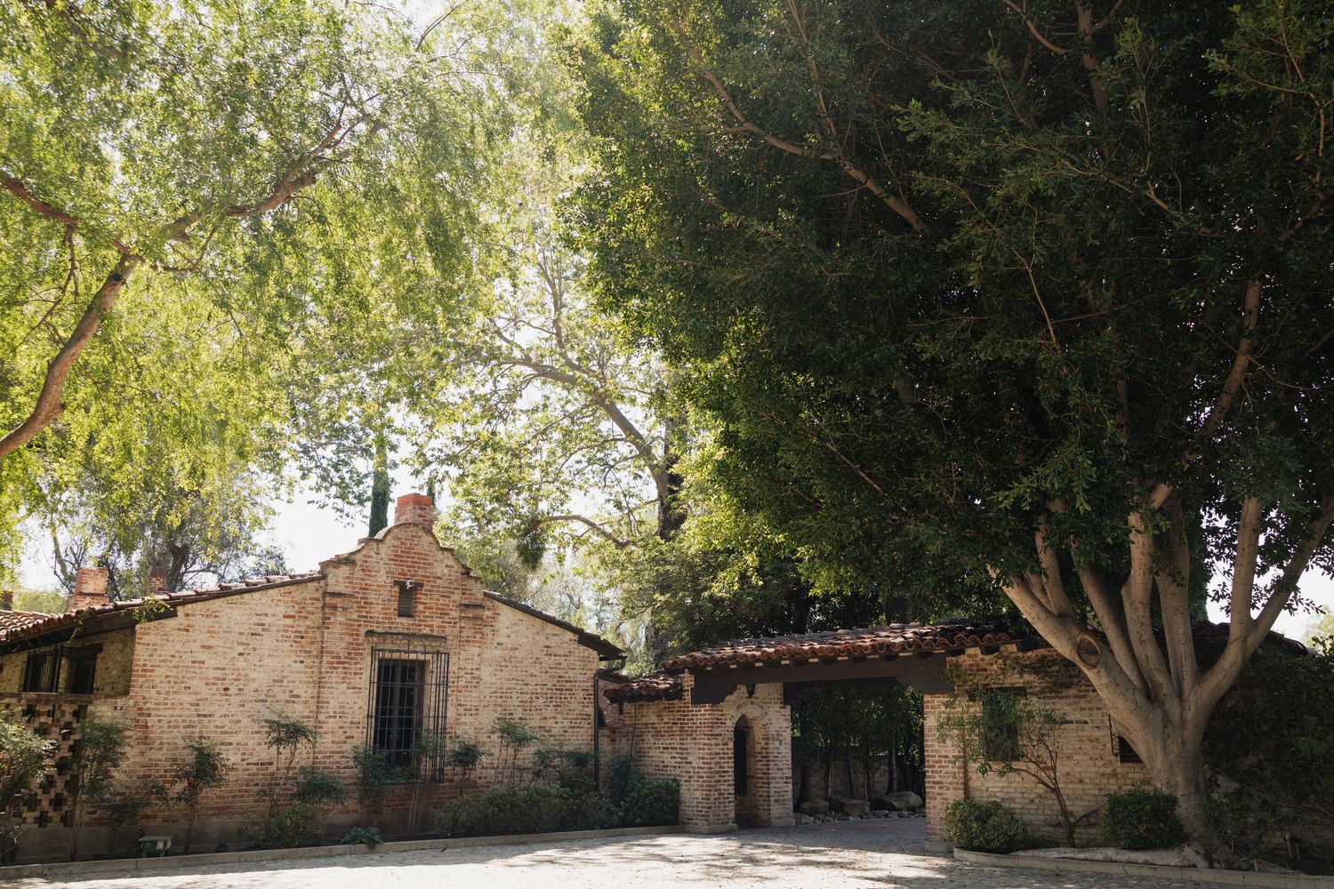 Rustic brick building surrounded by lush green trees on a sunny day, resembling a historic ranch or mission.