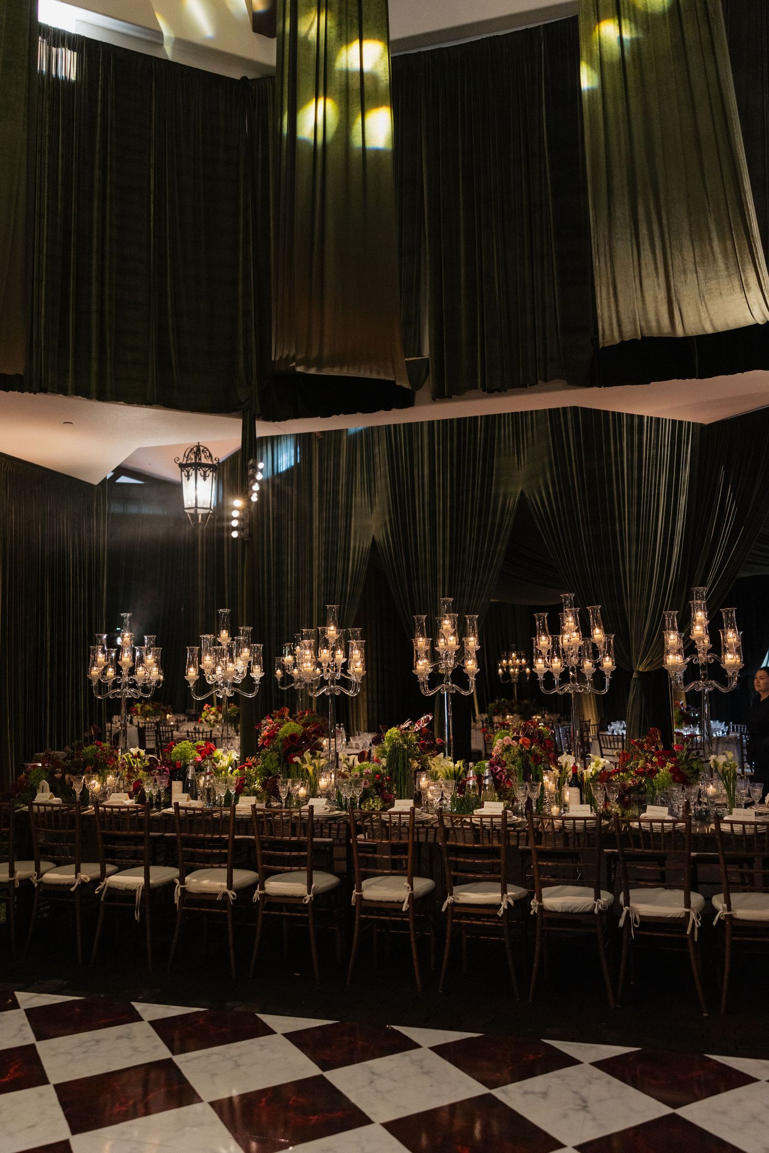 Dramatic wedding reception hall with dark draping, candelabras, colorful floral centerpieces, and checkered marble floor.