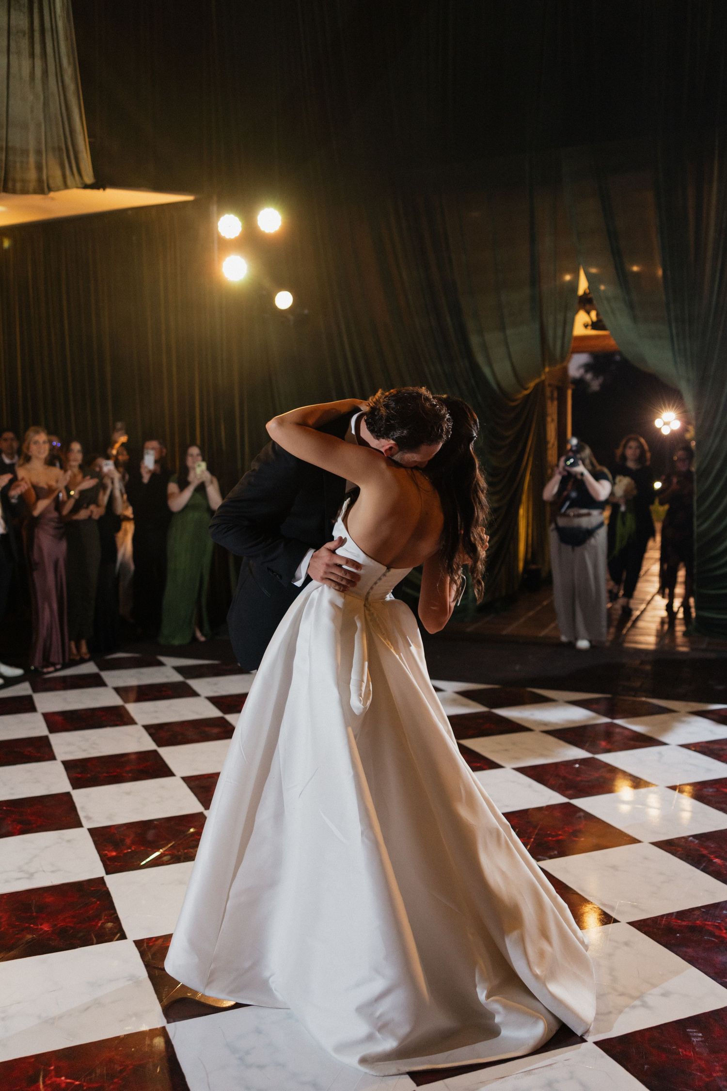 Bride and groom share first dance kiss on checkered floor, surrounded by applauding guests in elegant wedding venue.